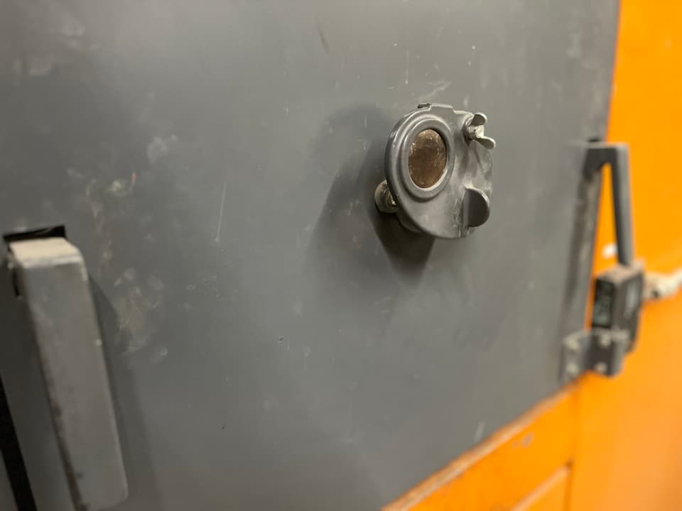 Nahaufnahme einer grauen Metalltür mit Türspion und orangefarbenem Hintergrund.