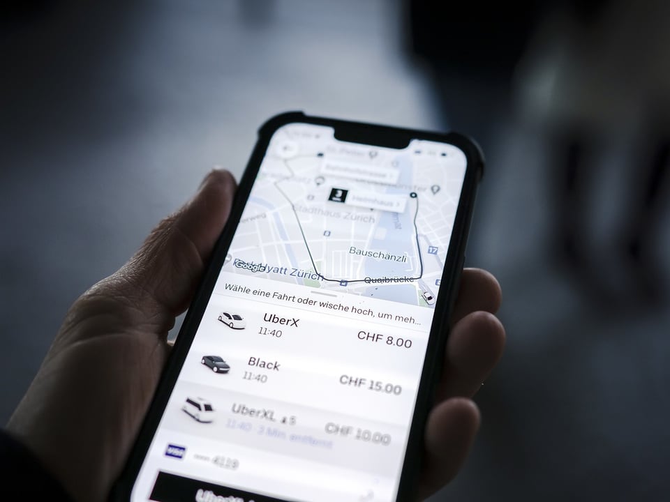 Ein Handy in einer Männerhand mit der App «Uber» offen, eine Landkarte mit einem schwarzen Punkt in der Mitte 