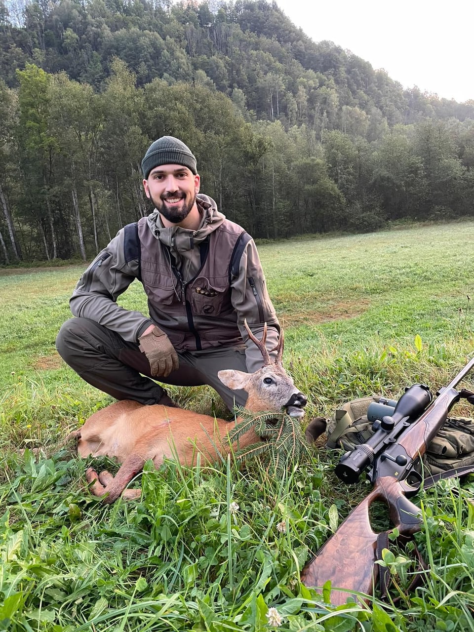 Jäger mit erlegtem Rehbock und Gewehr in einer Wiese.