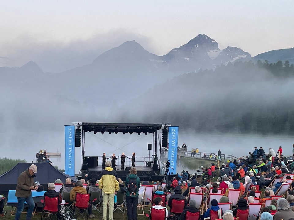 Konzertbühne vor nebelverhangenen Bergen, Zuschauer sitzen auf Stühlen im Freien.