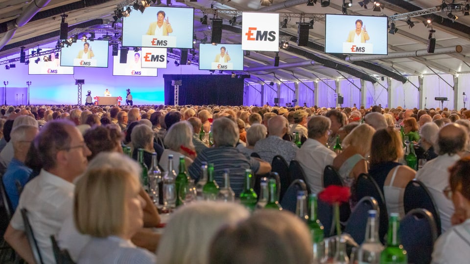 Grosses Publikum in einem Veranstaltungszelt mit mehreren Bildschirmen.