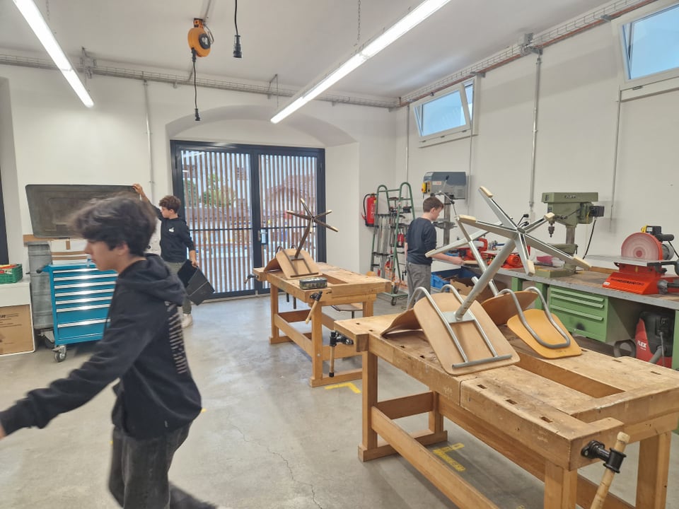 Es ist ein Werkraum mit Maschinen, Tischen und Stühlen zu sehen. Darin befinden sich drei Jugendliche.