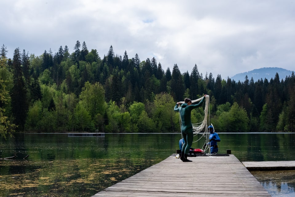 Menschen auf einem Steg an einem See mit Wald im Hintergrund.