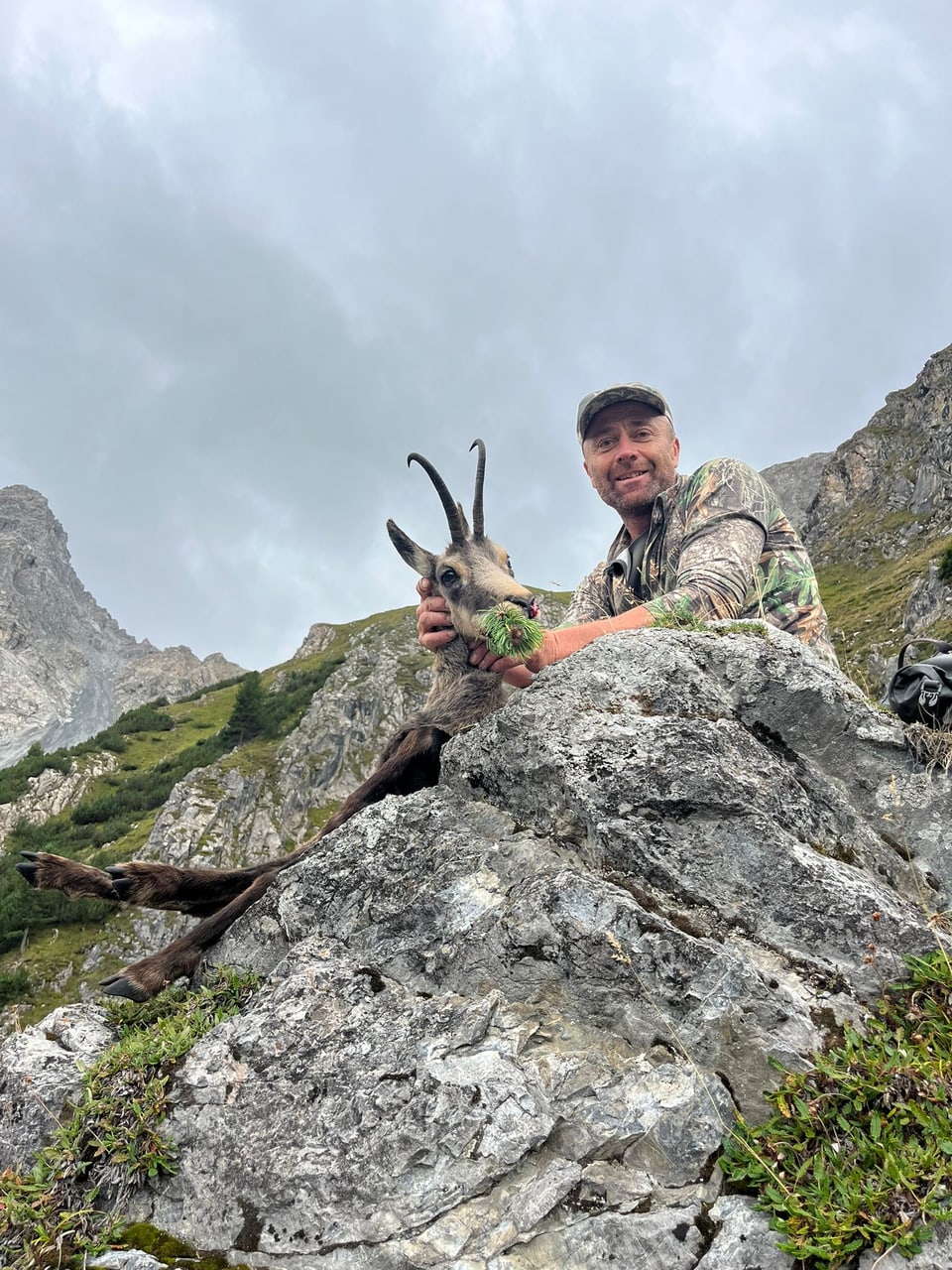 Mann posiert mit einem erlegten Steinbock in einer Berglandschaft.